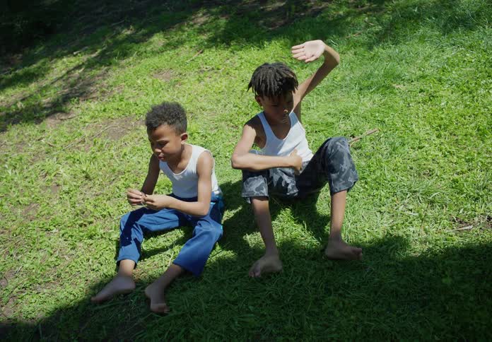 Two young boys sit together on the grassy while the sun shines brightly
