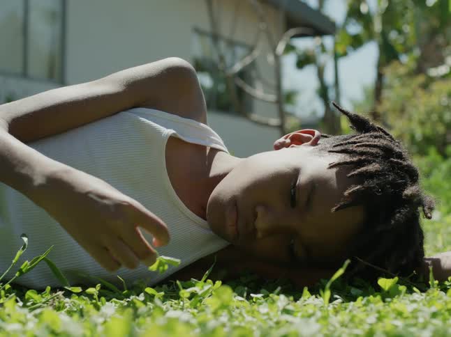 A boy idly plays with the grass while lying on the ground outdoors during the day