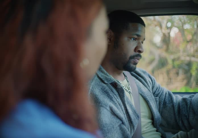 Man with necklace and gray jacket and woman with red curly hair and blue shirt sit in car quarrelling