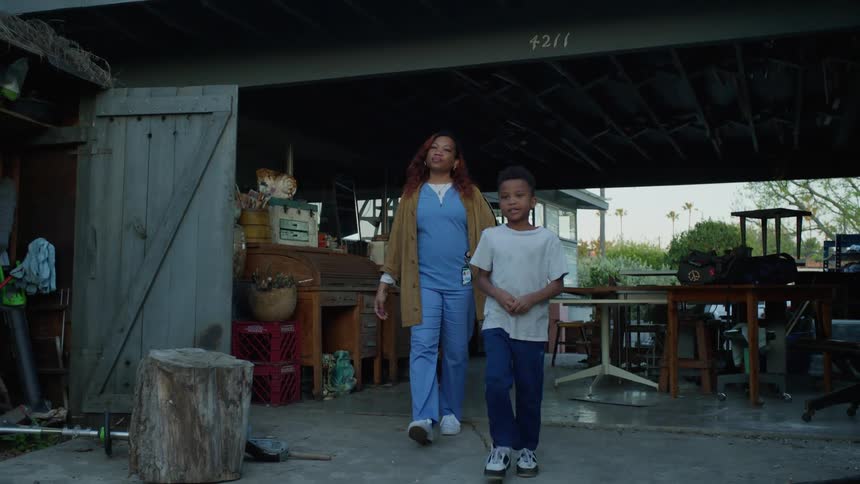 A mother wearing nurses scrubs walks out of a doorway with her two young sons into a room with furniture stacked all over the place in disarray