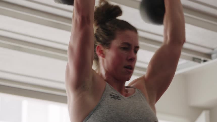 A very muscular female bodybuilder is in a gym doing kettlebell repetitions during the day
