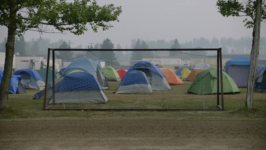 A large grassy area behind a goal net contains several tents and some rvs in the background
