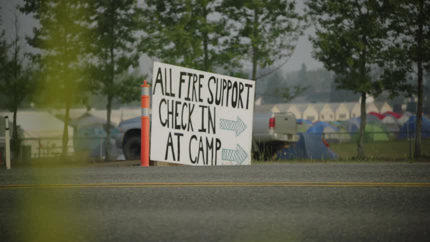A handmade sign by a road directs fire support professionals to check in at a camp of tents and buildings