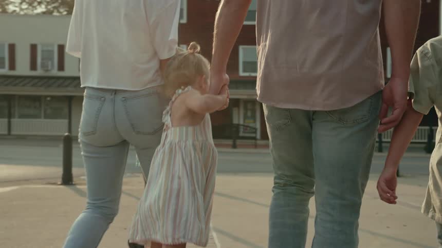 Little girl jumps while walking holding hands with her parents on the town streets at evening
