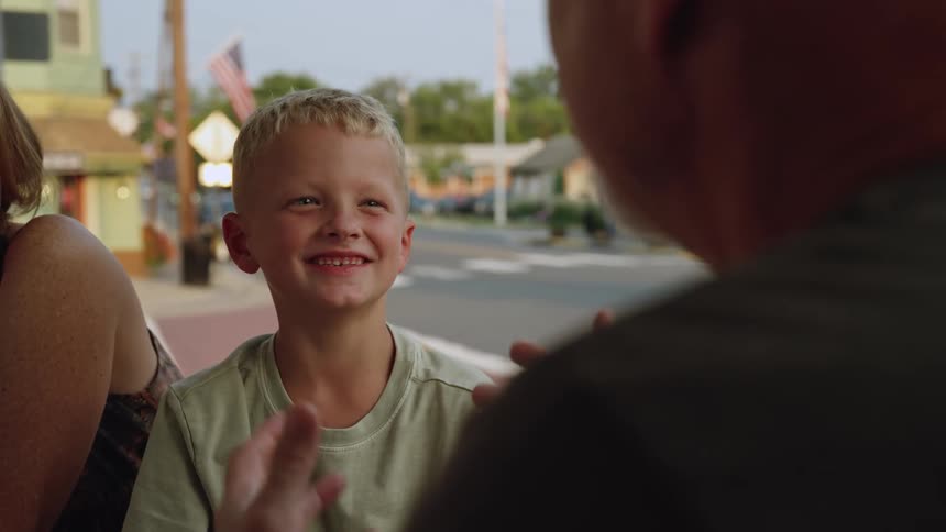Man using hand gestures talks to blonde boy in green shirt laughing at cafe