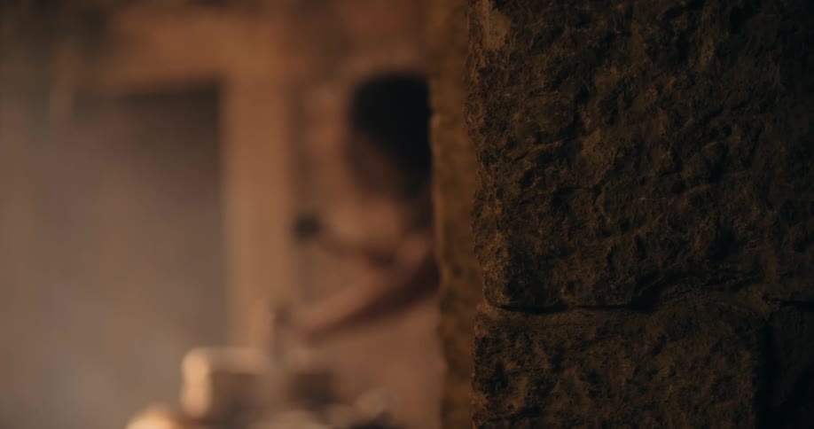 A man whittles a brick of wood behind a pillar in a dungeonlike setting