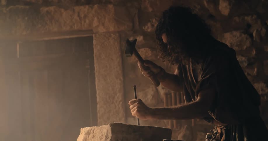 A sculptor uses a chisel and hammer to hit a large rock starting a new artwork in a room with stone walls