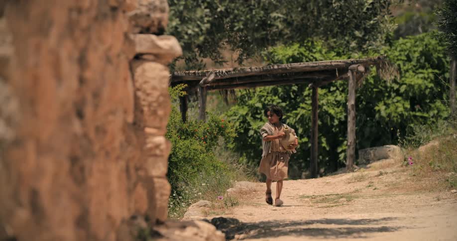A young boy carrying a clay jug runs along a dirt path next to a building