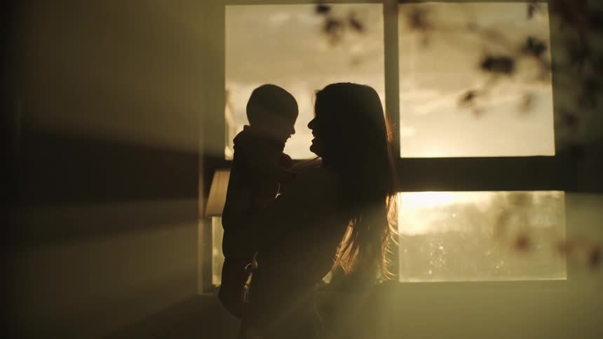 Mother with long brown hair holding toddler son is silhouetted against the sun shining through windows