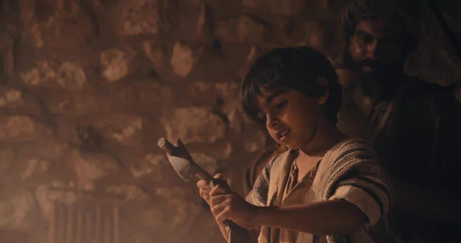 A young boy uses a hammer and chisel as an adult nearby drinks water from a jug