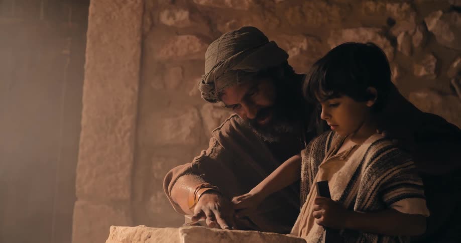 A man shows a young boy how to carve into stone using a sharp blade and a hammer
