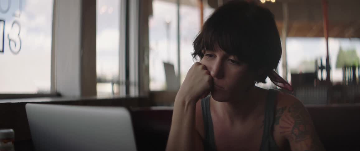 Tattooed woman sitting at the table looks at a laptop and sighs at a cafe during daylight