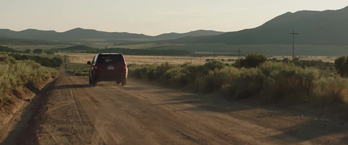 A red suv travels on a dirt road with brush road side and hills on the horizon