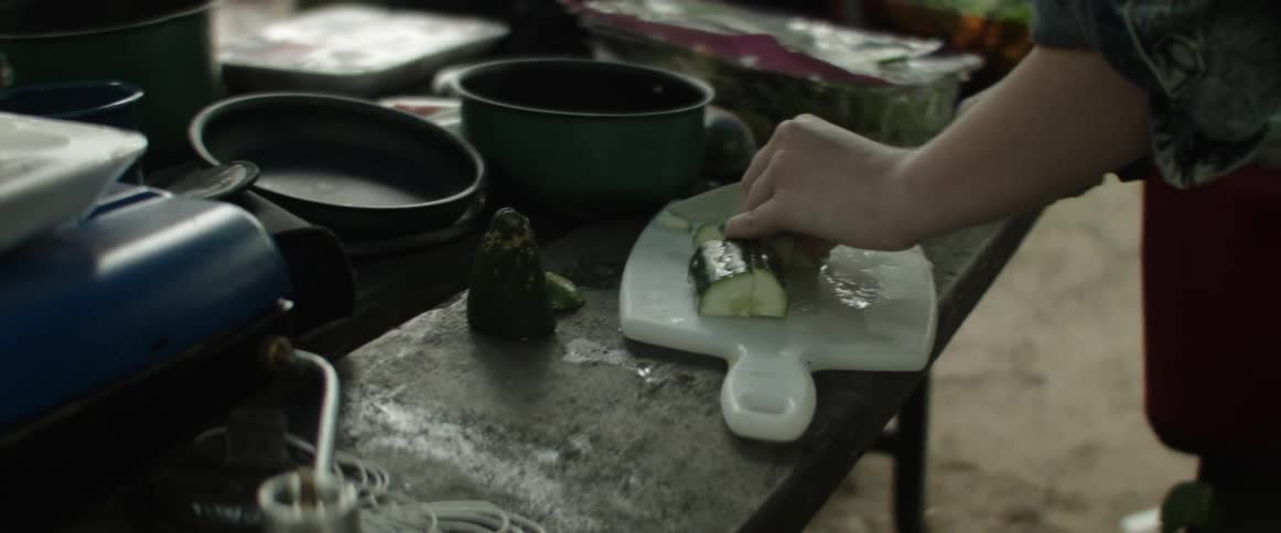 A woman stands at a campsite in the forest cutting a cucumber on a cutting board wearing a baseball hat
