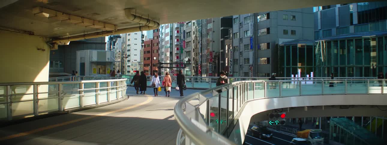 People with shopping bags walk on a large pedestrian bridge that connects buildings in a city