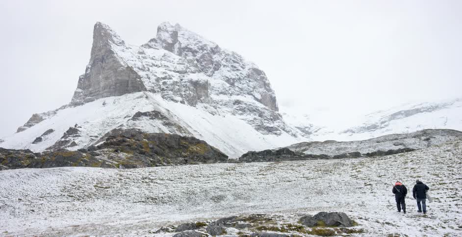Two people walk toward a craggy peak in a snowy landscape