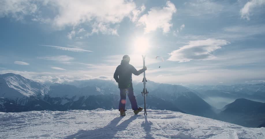 The sun shines brightly upon a man standing on a mountain holding his skis as he watches a paraglider fly over mountains in the distance
