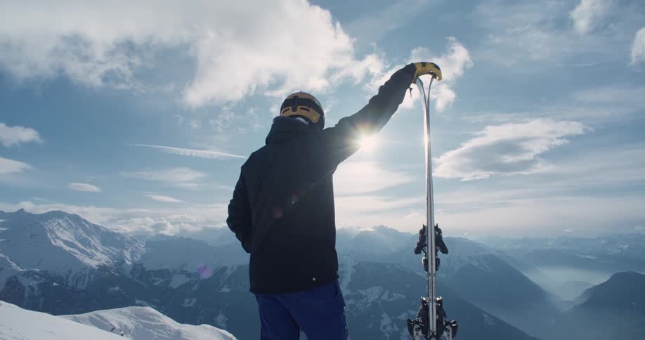 Skier stands with one hand on skis sitting upright on ground and looks over snow covered mountains on a sunny day