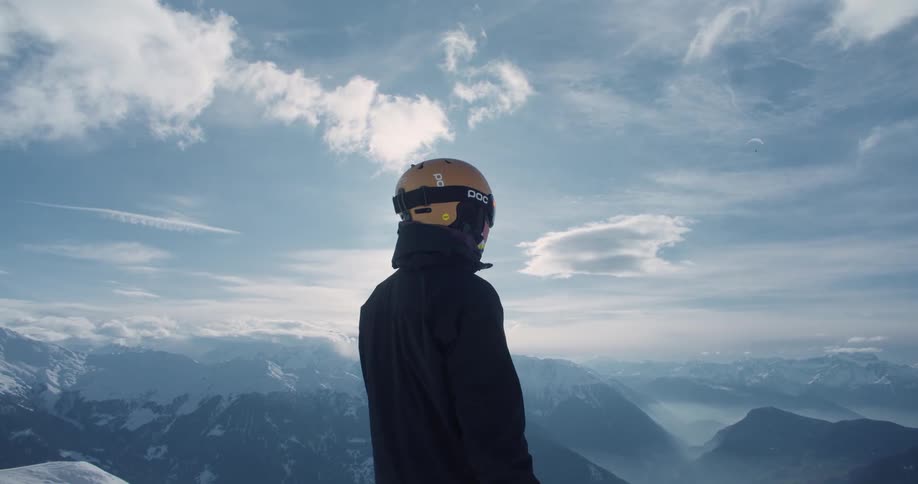 The sun shines brightly upon a man wearing a helmet and standing on a mountain looking at snow covered mountains in the distance