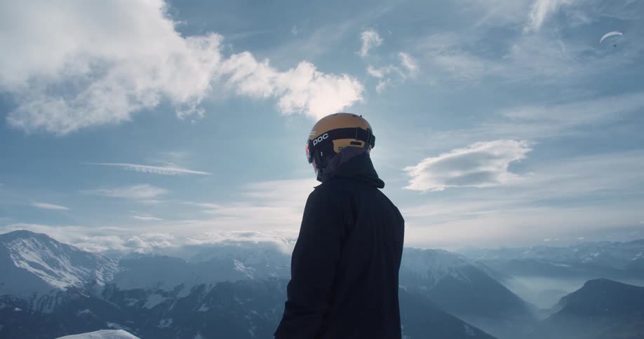 The sun shines upon a man wearing a helmet standing on a mountain as he looks at snow covered mountains in the distance