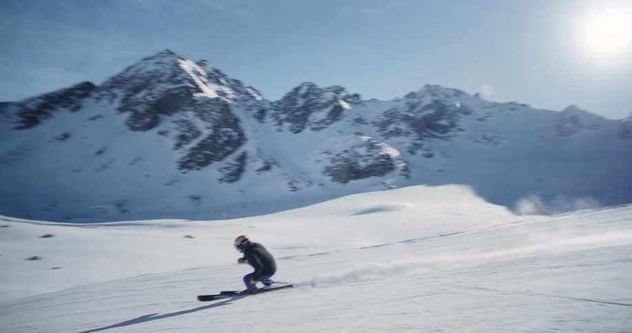 The sun shines brightly upon a skier as he holds his poles and zigzags down the snow covered slope and there are snow covered mountains in the distance