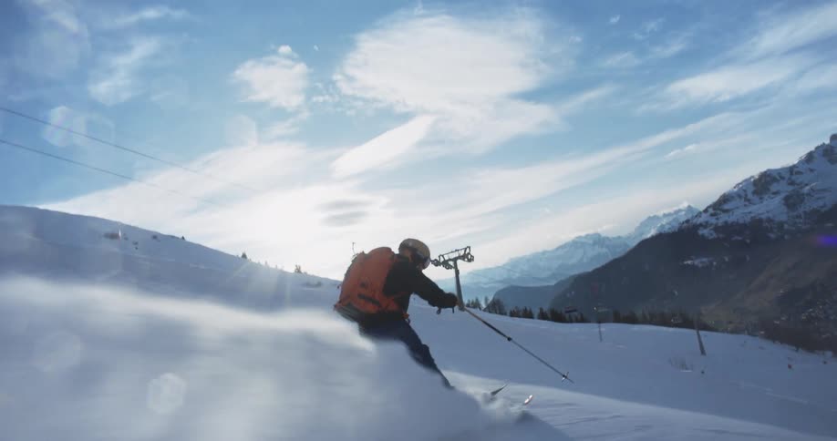 A skier wearing a backpack holds the ski poles and makes his way down the hill as snow flies behind him