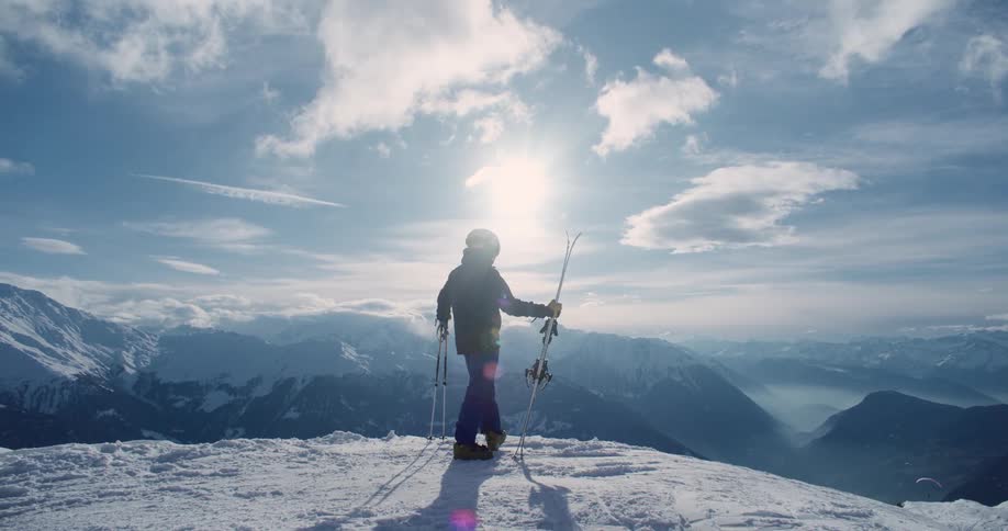 Person standing at top of snow covered mountain holding skis and poles stopped and admiring view