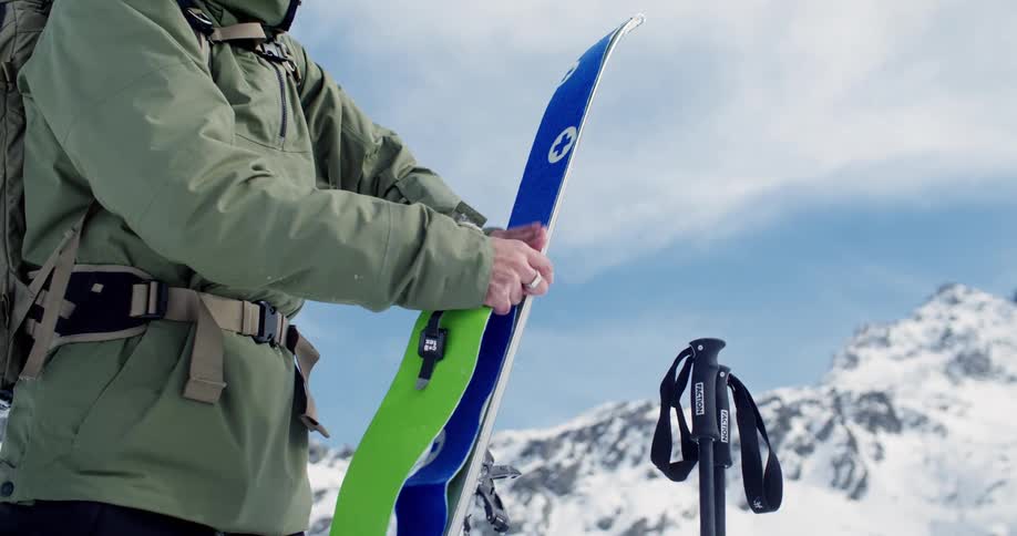 A man at the bottom of ski run placing ski skins on skis in preparation with snowcovered peaks behind him