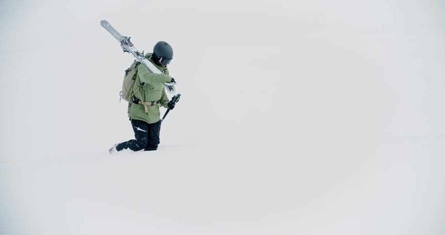 A mountaineer walks through a field of deep snow while carrying skis over one shoulder