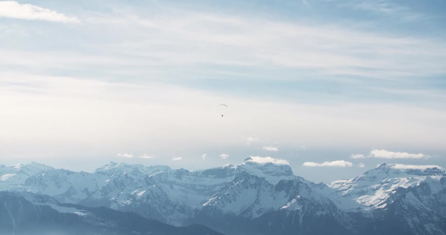 Panoramic view of a person parachute gliding over a mountain valley snowy mountain range and a cloudy sky is seen in the distance