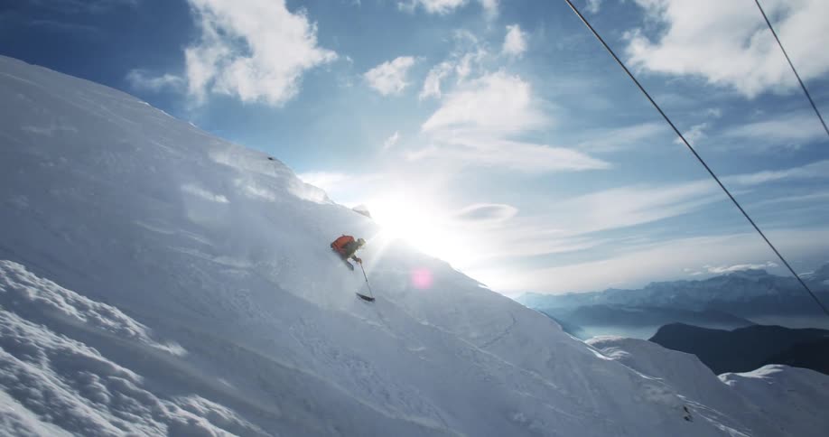 A skier holds his poles and zigzags down the snow covered slope moving under ski lifts as the sun shines over the snow covered mountain