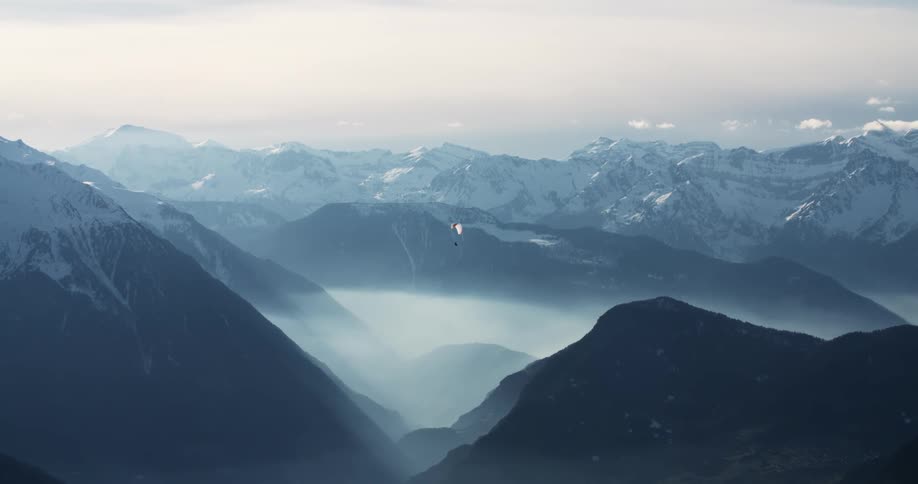 Paraglider floats through air over dense foggy valley surrounded by snow topped mountains