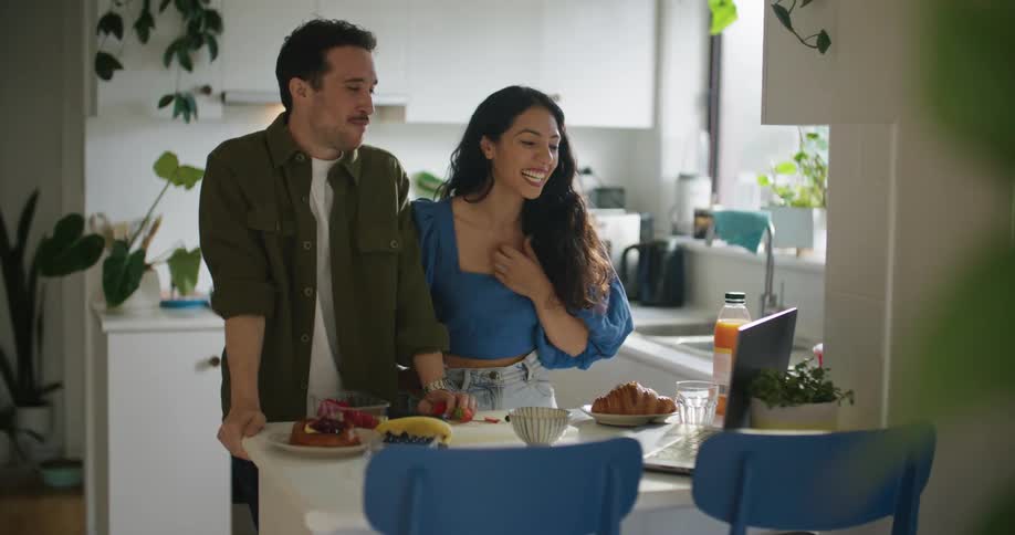 Man with mustache green shirt and woman with wavy hair blue shirt and jeans make video call on laptop in the kitchen laughing