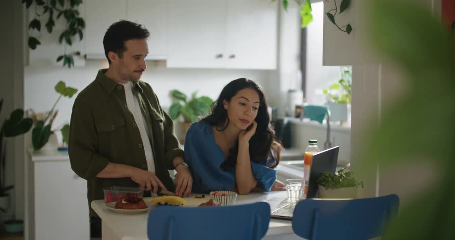 Man and woman at breakfast bar in kitchen at home looking at home decor on the laptop and eating breakfast