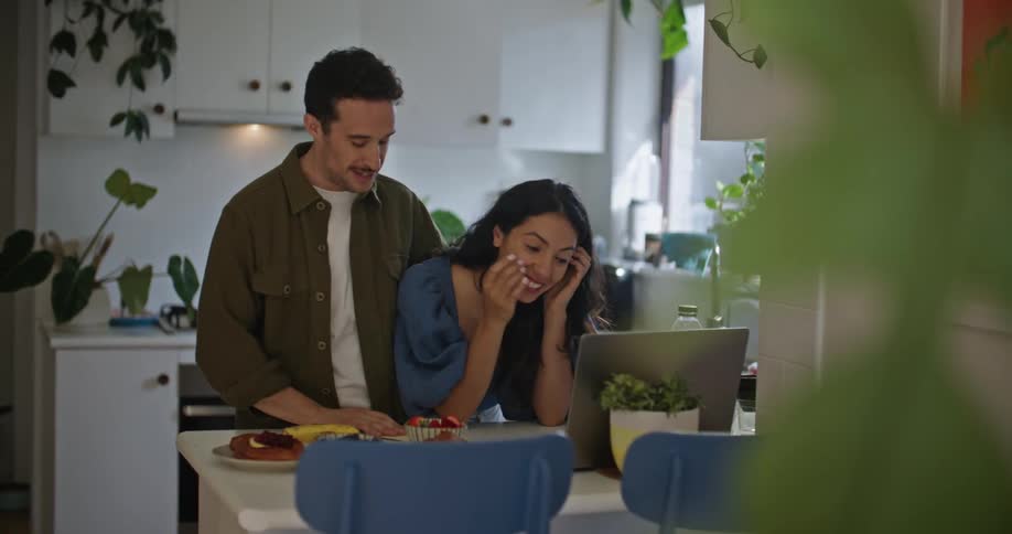 A man and a woman at their kitchen table talk to someone through a laptop