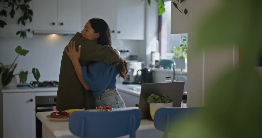 Man in yellow shirt and woman wearing blue shirt and jeans cuddle in front of laptop in the kitchen