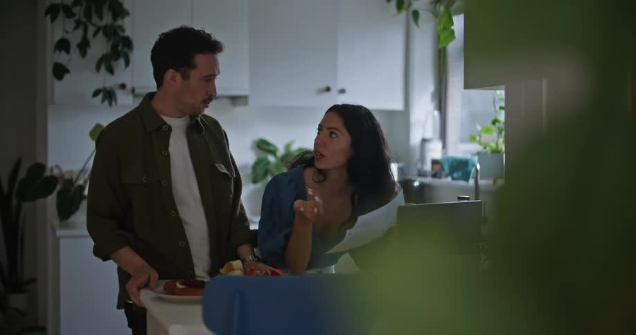 Man with mustache green shirt and woman with dark hair and blue shirt read paper in front of laptop in the kitchen