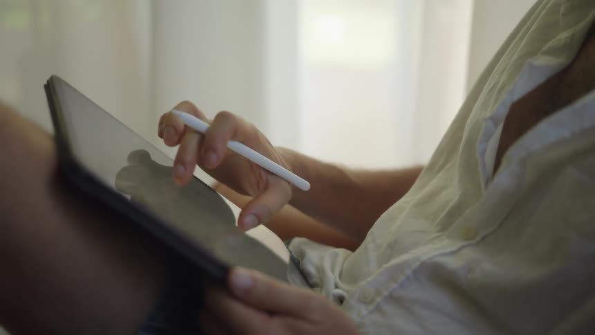 A man is sitting down holding a tablet computer in his lap as he writes on the screen with a stylus