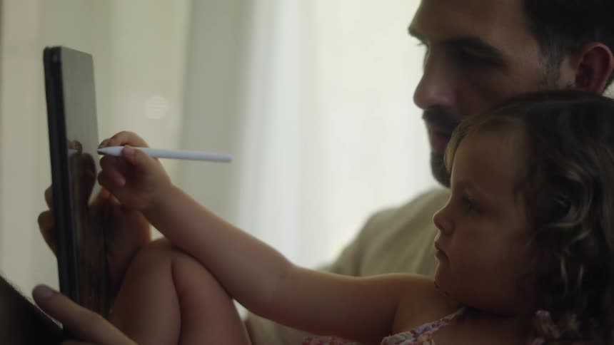 A girl uses a stylus on a computer tablet while being cradled in the arms of a man sitting indoors during the day