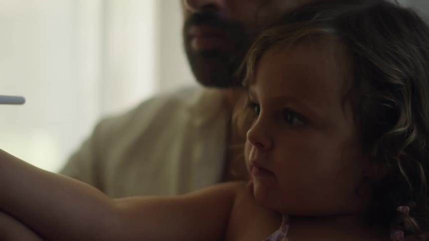 A little girl sitting on her fathers lap while she draws using a white pencil