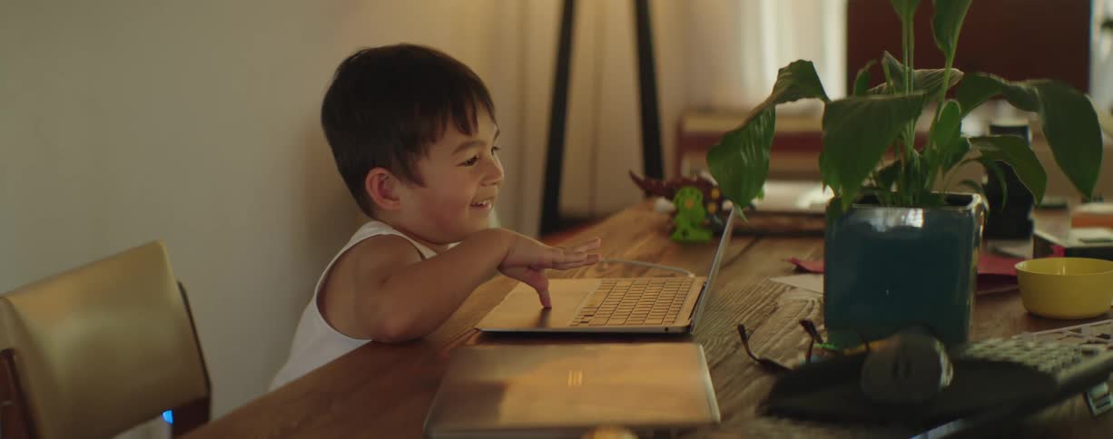 A little boy is sitting at a table laughing as he plays on a laptop computer