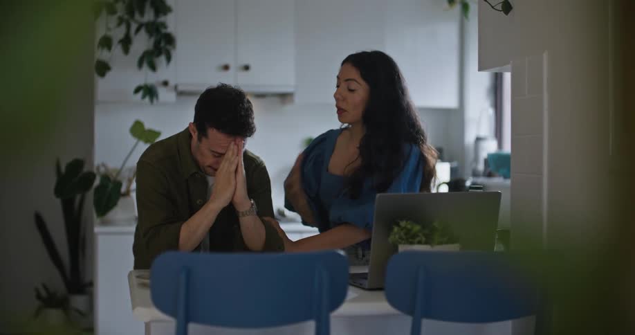 A man and a woman are at the kitchen table with a laptop computer as they talk and hug