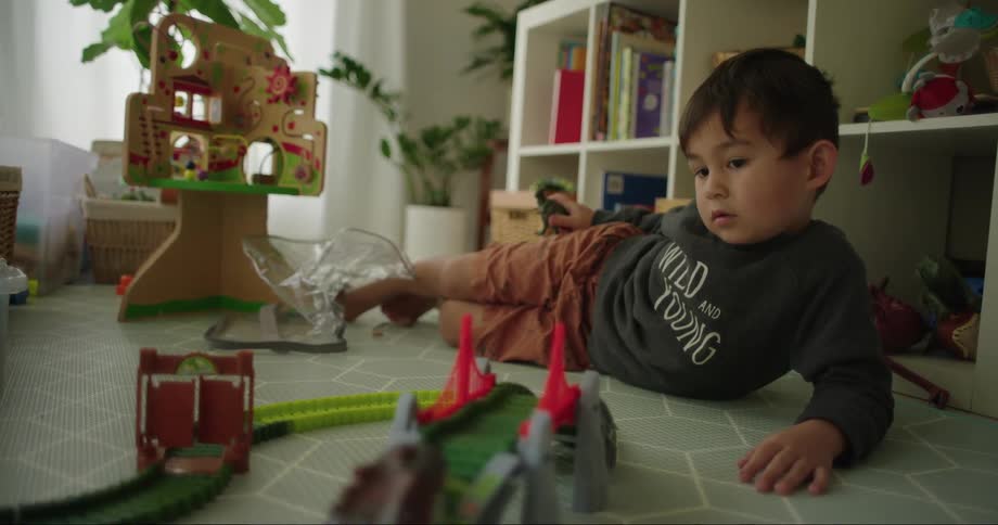 A toddler boy is laying on the floor and watching a toy car go around a track