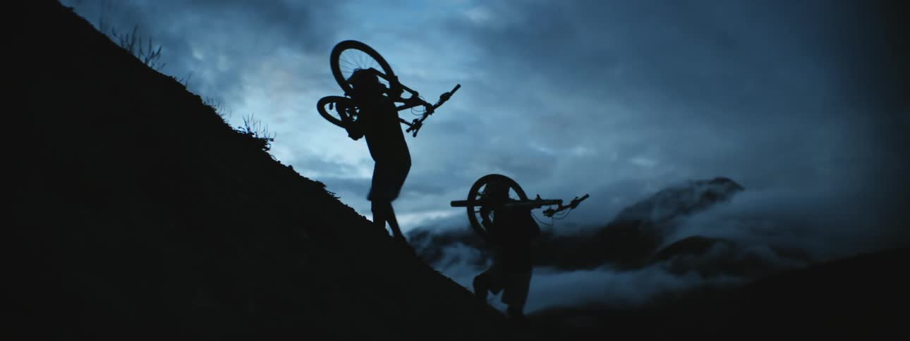 A pair of cyclists walk up a steep hill while carrying their bicycles on their shoulders