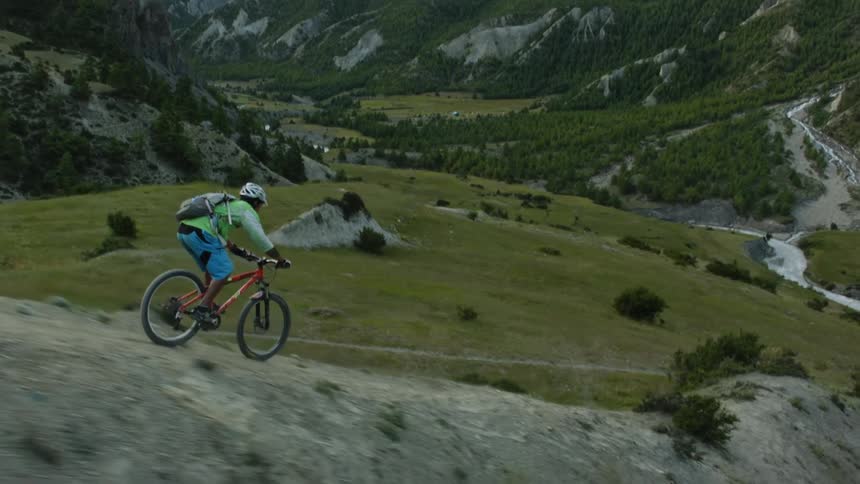 Two men on bicycles ride down a slope toward a forested valley before mountain peaks