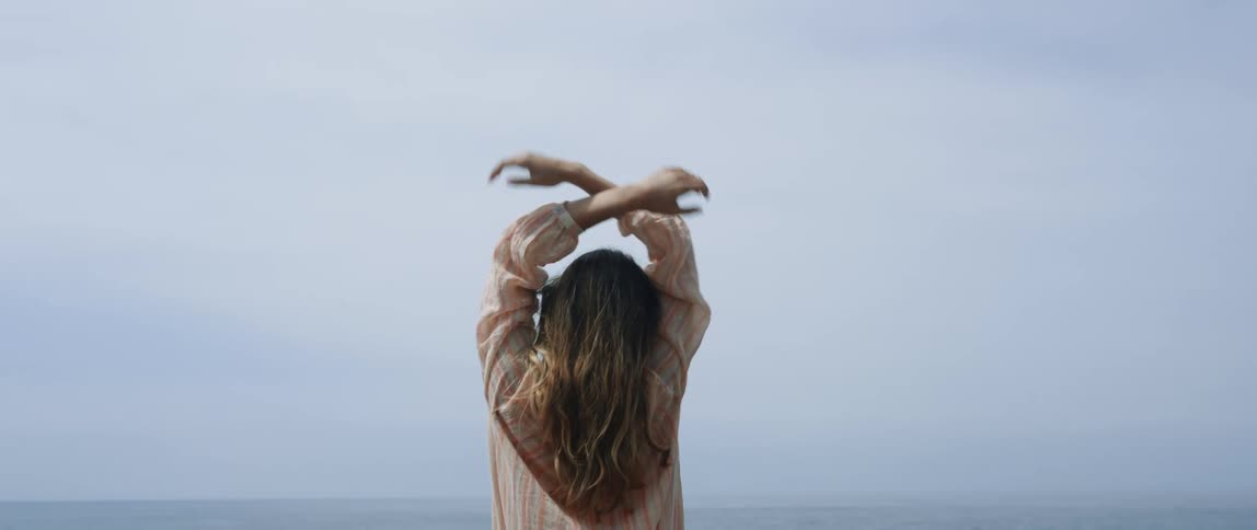 Woman with brown hair and shirt raises her arms up and watches ocean