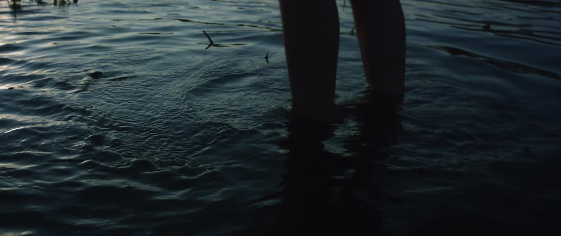 Person standing in shallow part of a lake with reeds in it swirling feet around and making small waves reflection of sunset on the water