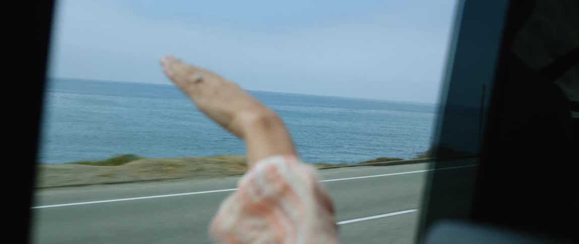 Hand of a woman out a moving car window waving in the wind in front of the ocean