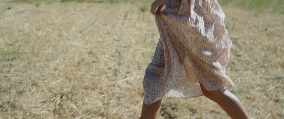 A woman wearing a print dress and sandals running on a farm field in the sunshine