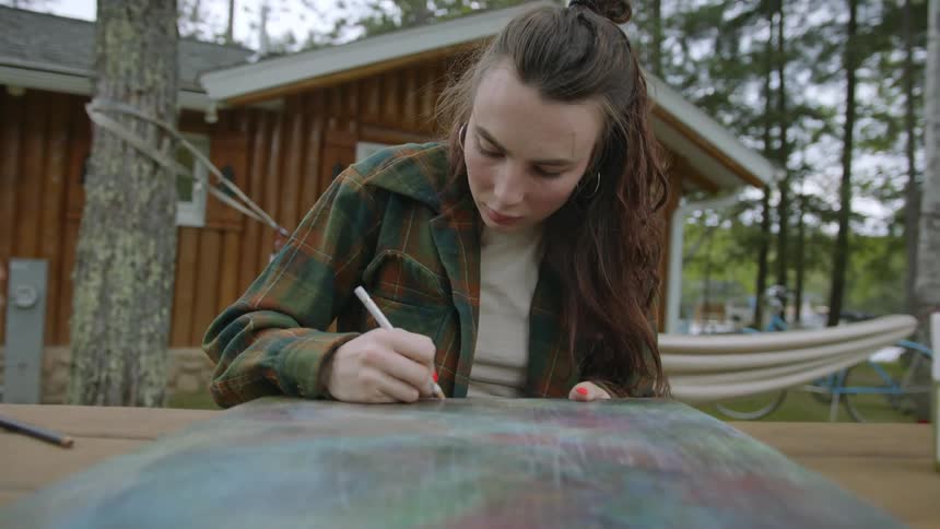 A woman sitting at a table outside while she draws on a canvas